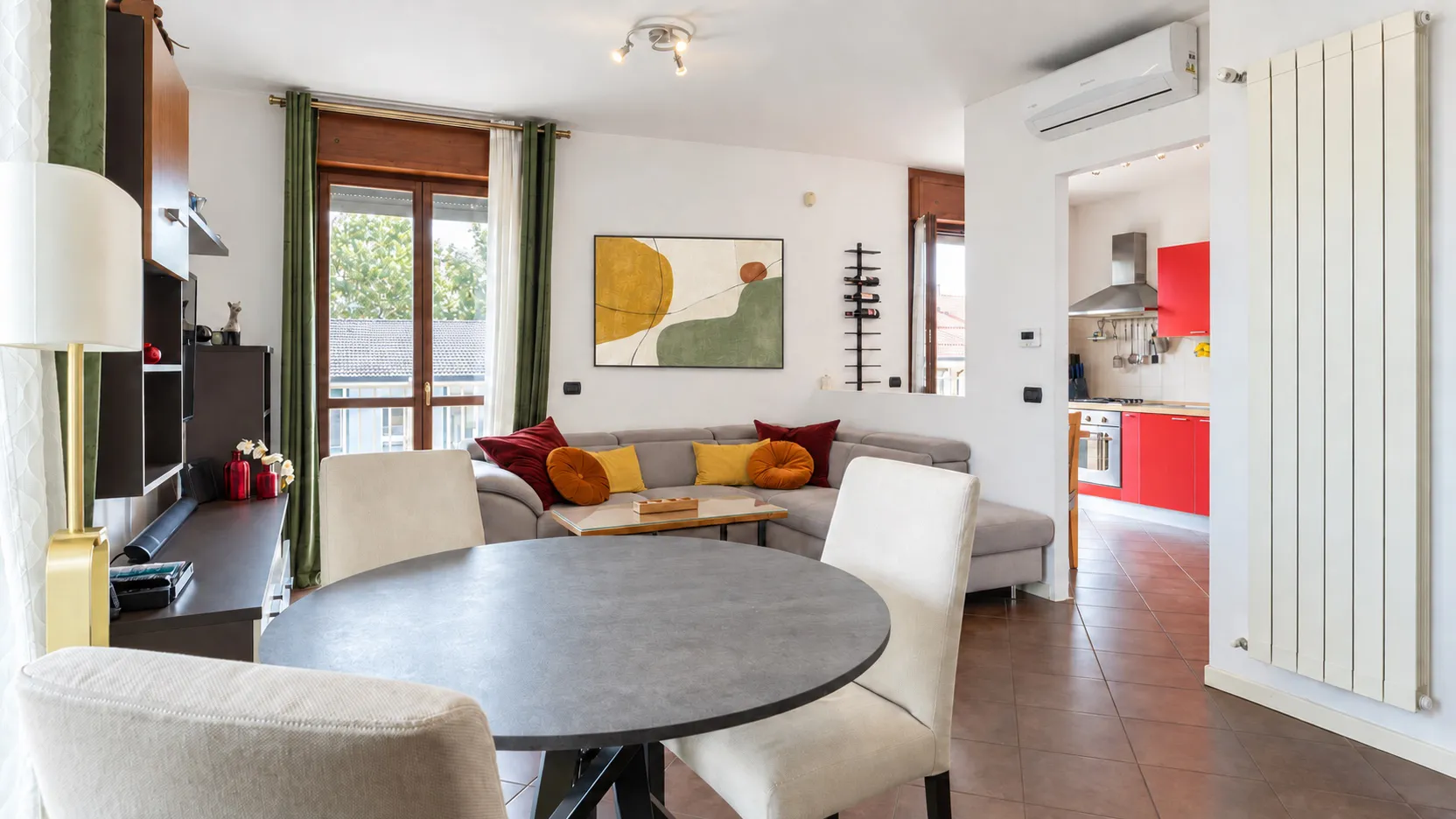Zona pranzo con il tavolo rotondo scuro e le sedie chiare, il divano sullo sfondo e, oltre il passaggio, la cucina rossa